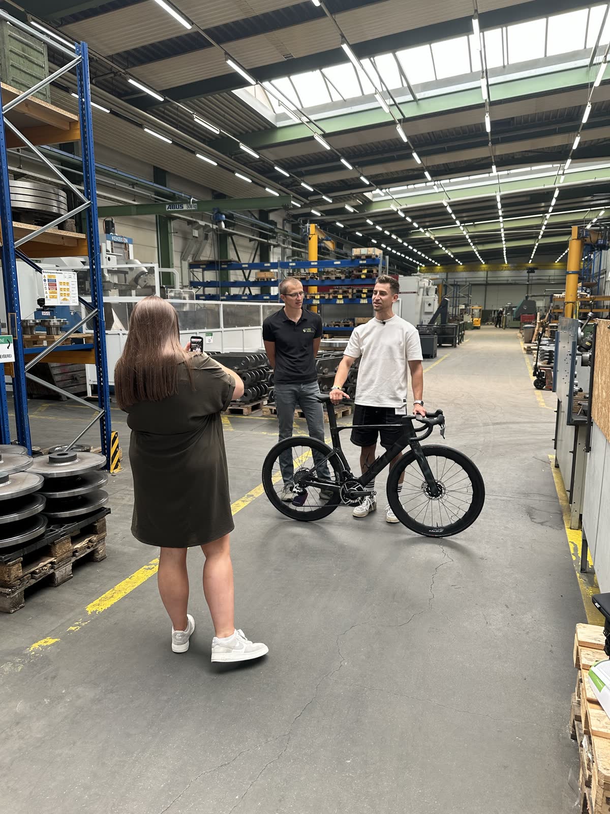 Dreh in einer Werkshalle: Mitarbeiterin filmt zwei Kollegen mit Rennrad